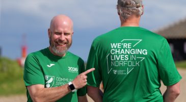 one man in green top pointing at the back of another mans top that says changing lives in norfolk